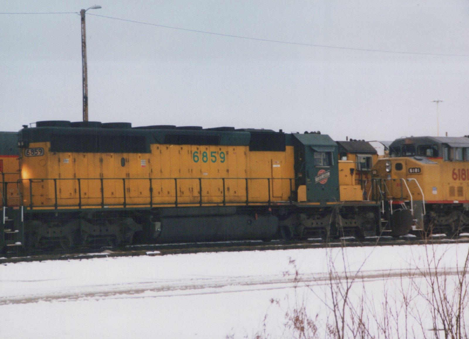 A yellow and green Chicago and North Western diesel locomotive, number 6859, sits on tracks in a snowy yard, viewed from the side, with another locomotive behind it.