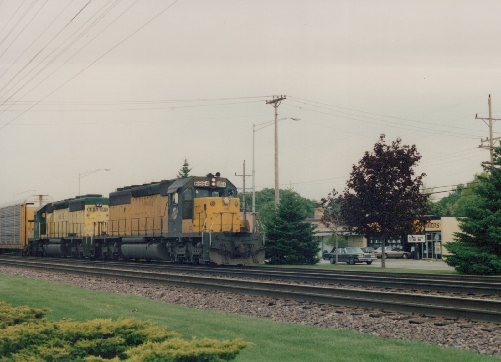 A yellow and green diesel locomotive with the number 6864 leads a train, followed by a second locomotive, on a set of tracks beside a road with parked cars and trees.