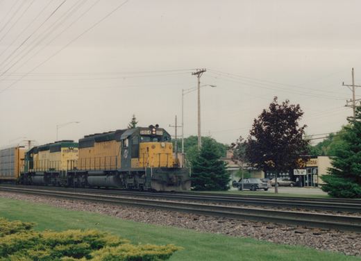 A yellow and green diesel locomotive with the number 6864 leads a train, followed by a second locomotive, on a set of tracks beside a road with parked cars and trees.