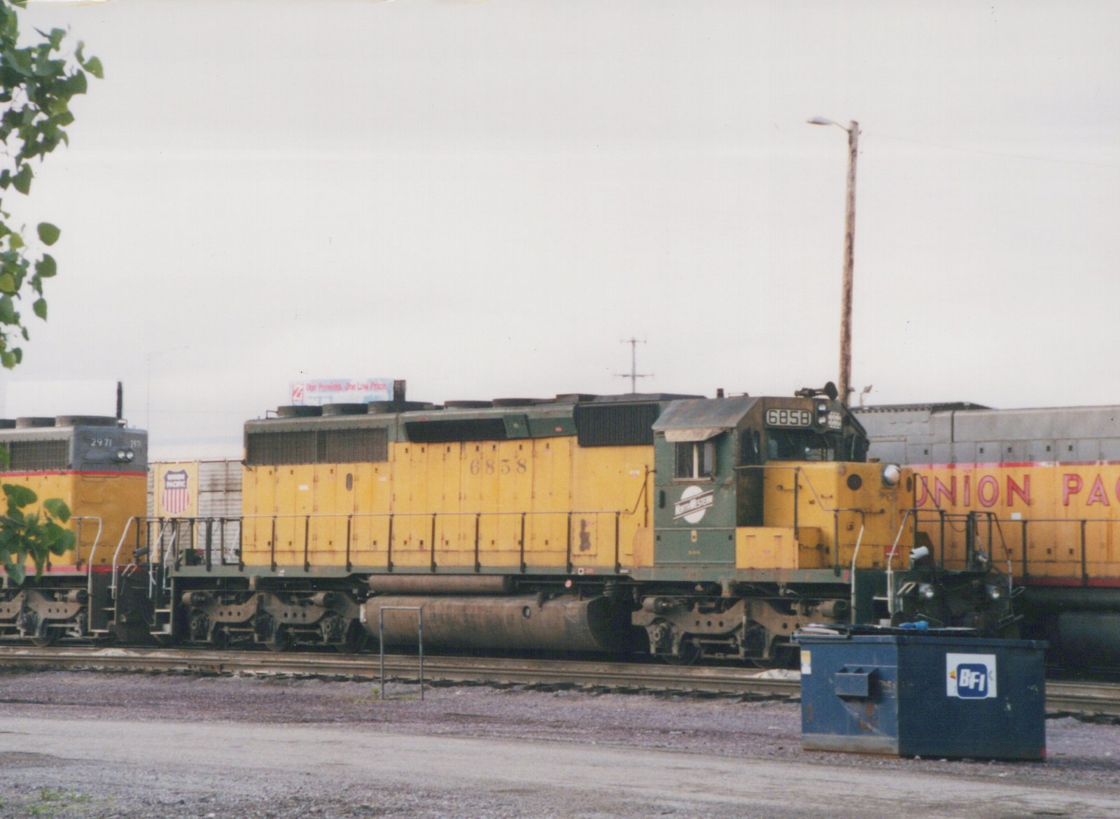 A yellow and green Chicago and North Western locomotive numbered 6858 sits on tracks in a rail yard, with a yellow Union Pacific locomotive visible behind it.