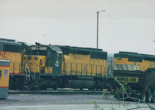A yellow and green Union Pacific diesel locomotive numbered 6858 is parked in a railroad yard on an overcast winter day, with other locomotives and a truck carrying an oversize load nearby.