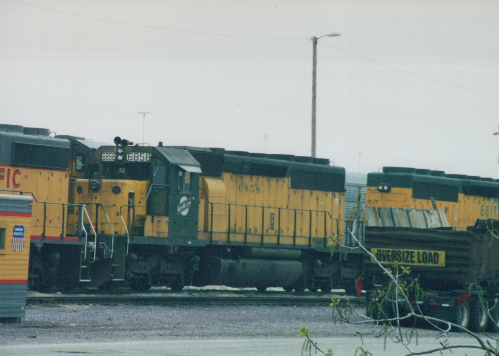 A yellow and green Union Pacific diesel locomotive numbered 6858 is parked in a railroad yard on an overcast winter day, with other locomotives and a truck carrying an oversize load nearby.