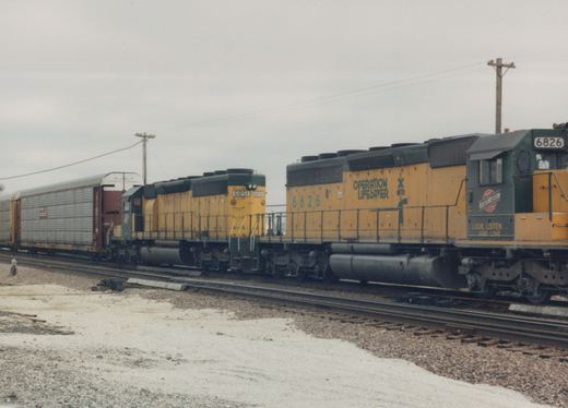 Two yellow and green Chicago and North Western GP9 diesel locomotives, numbered 6826 and 6851, are coupled together on a set of railroad tracks in a yard, with freight cars behind them and snow on the ground.