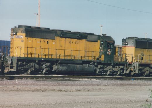 Side view of a yellow and green Chicago and North Western GP9 diesel locomotive, number 6847, in a gravel yard with another locomotive behind it.