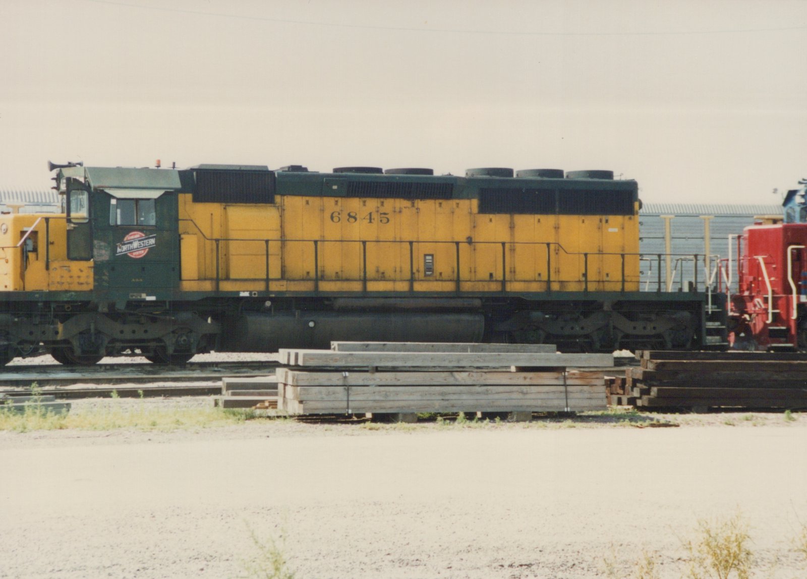 A side view of a yellow and green Chicago and North Western diesel locomotive, number 6845, sitting on a track next to stacks of wooden ties.