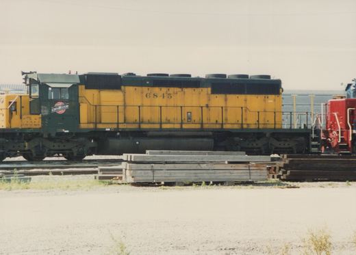 A side view of a yellow and green Chicago and North Western diesel locomotive, number 6845, sitting on a track next to stacks of wooden ties.