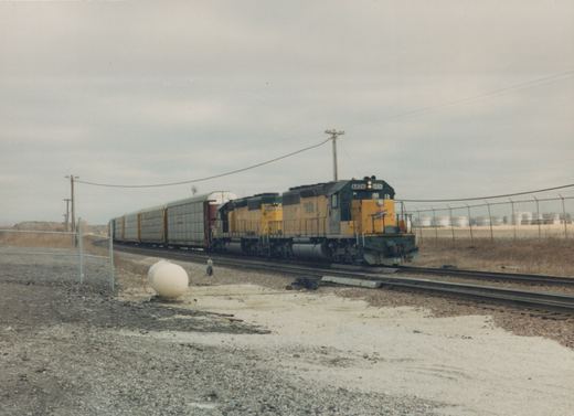 A yellow and green diesel locomotive, number 6826, pulls a line of freight cars on a railroad track, with a chain-link fence and industrial tanks in the background.