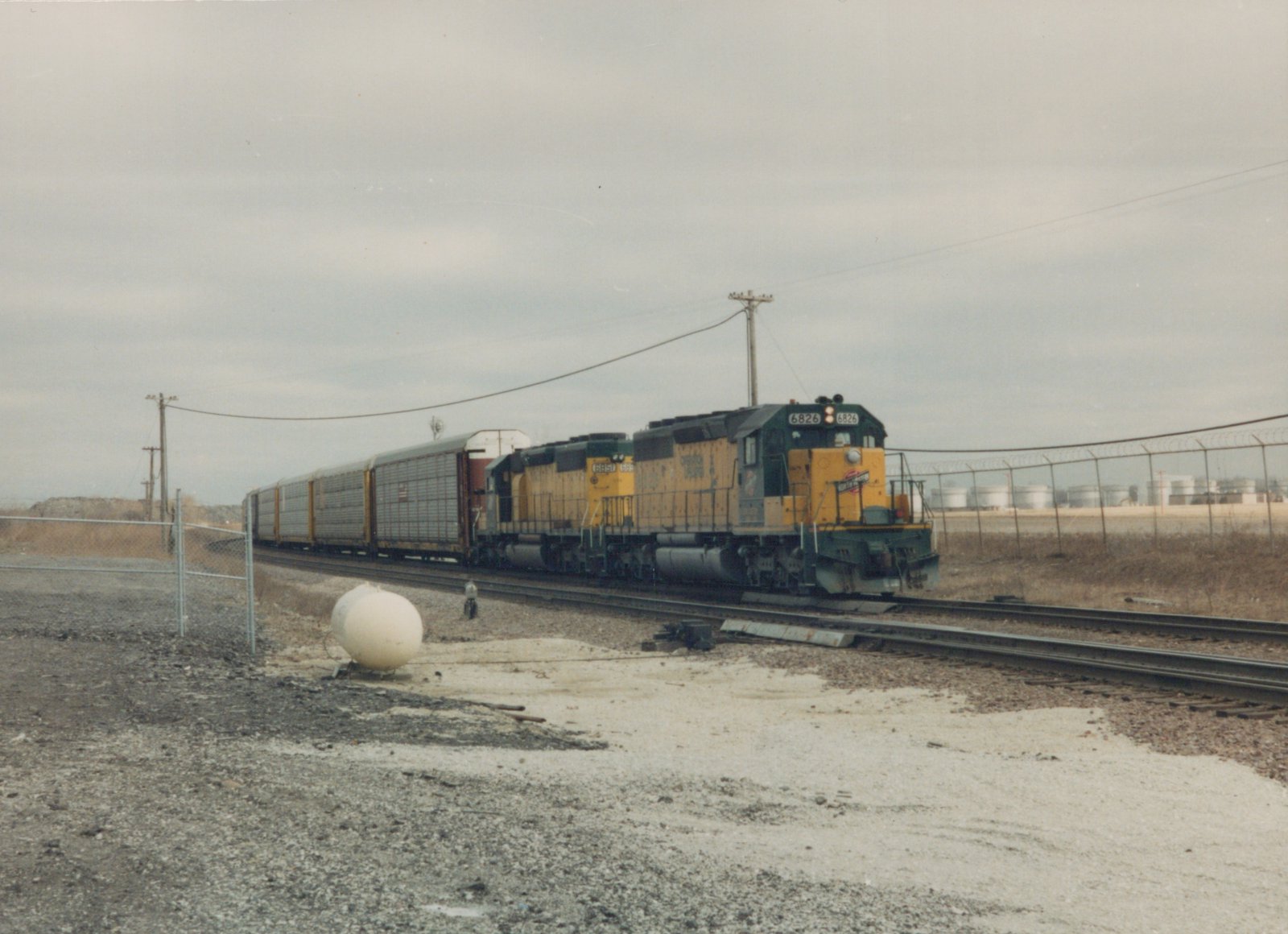 A yellow and green diesel locomotive, number 6826, pulls a line of freight cars on a railroad track, with a chain-link fence and industrial tanks in the background.