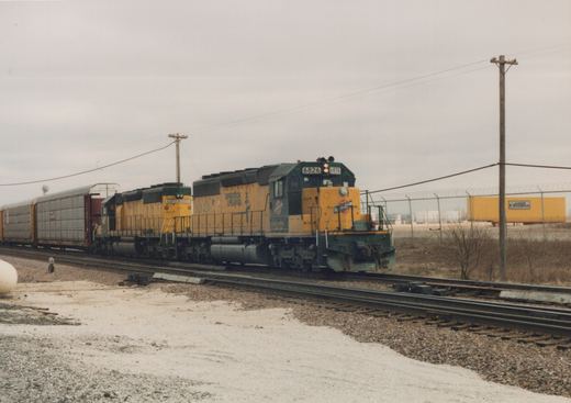 A yellow and green Chicago and North Western diesel locomotive numbered 6826, with 'Operation Lifesaver' lettering, leads a short train of freight cars on a snowy day.