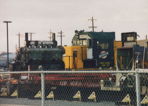 A yellow and black diesel locomotive with 'Chicago and North Western' lettering and the number 6825 is parked in a rail yard behind a chain-link fence, with a red flatcar and a large green engine block nearby.