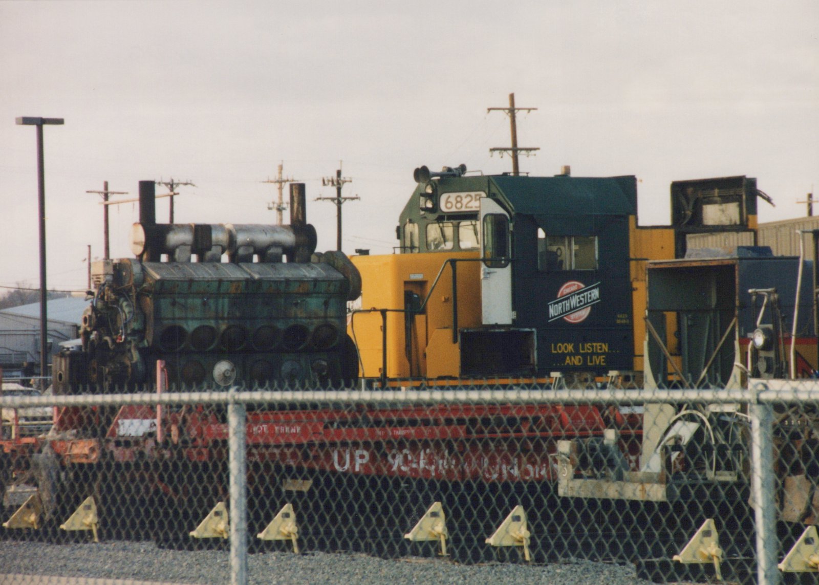 A yellow and black diesel locomotive with 'Chicago and North Western' lettering and the number 6825 is parked in a rail yard behind a chain-link fence, with a red flatcar and a large green engine block nearby.