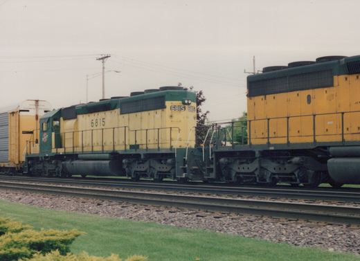 A yellow and green diesel locomotive with the number 6815 and CNW lettering is shown on train tracks beside a grassy area under an overcast sky.