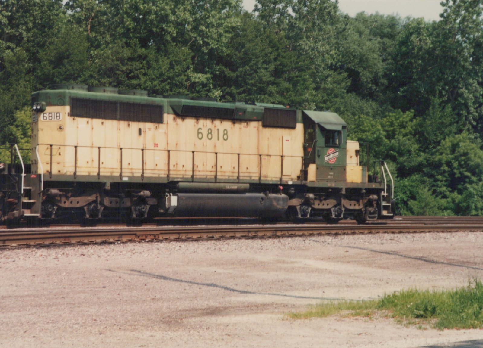 A green and yellow Chicago and North Western diesel locomotive, number 6818, sits on tracks in a gravel yard with trees in the background.