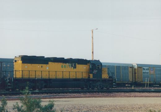 A side view of a yellow and black Chicago and North Western SD24 locomotive, numbered 6811, pulling a freight car on a railroad main line under an overcast sky.