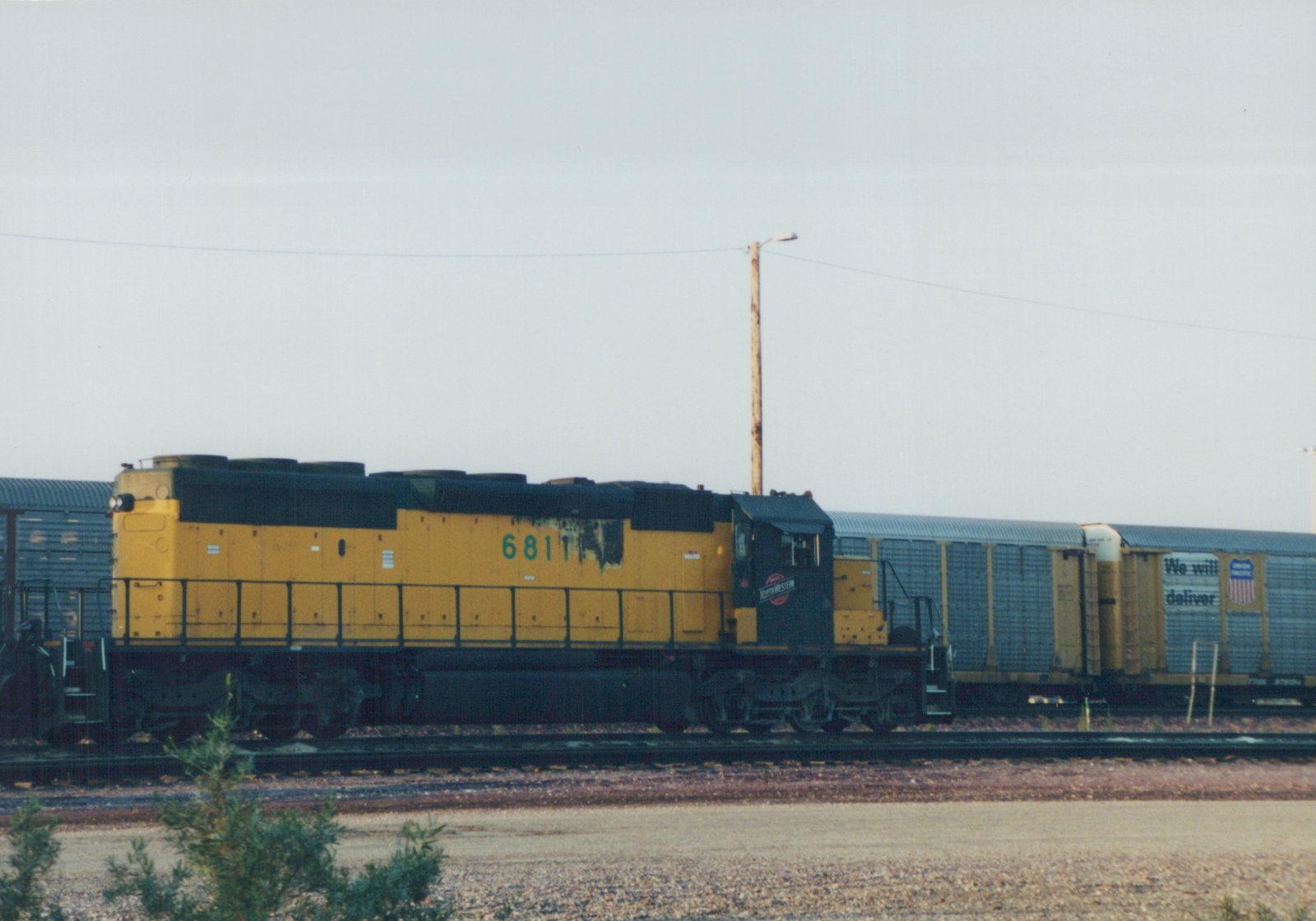 A side view of a yellow and black Chicago and North Western SD24 locomotive, numbered 6811, pulling a freight car on a railroad main line under an overcast sky.