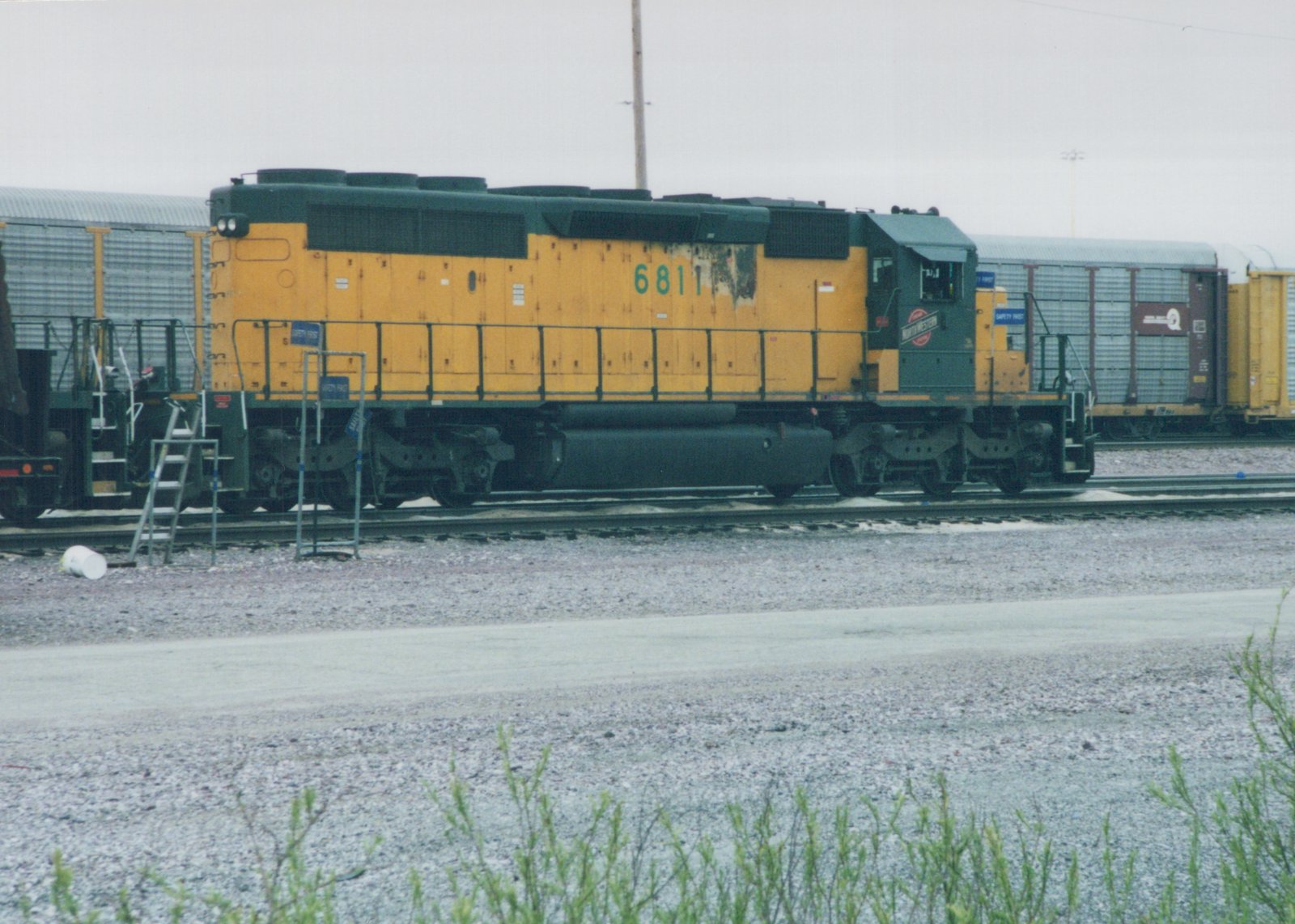 A yellow and green diesel locomotive with the number 6811 and a Northwestern herald on the cab, parked on tracks in a gravel yard with freight cars in the background.
