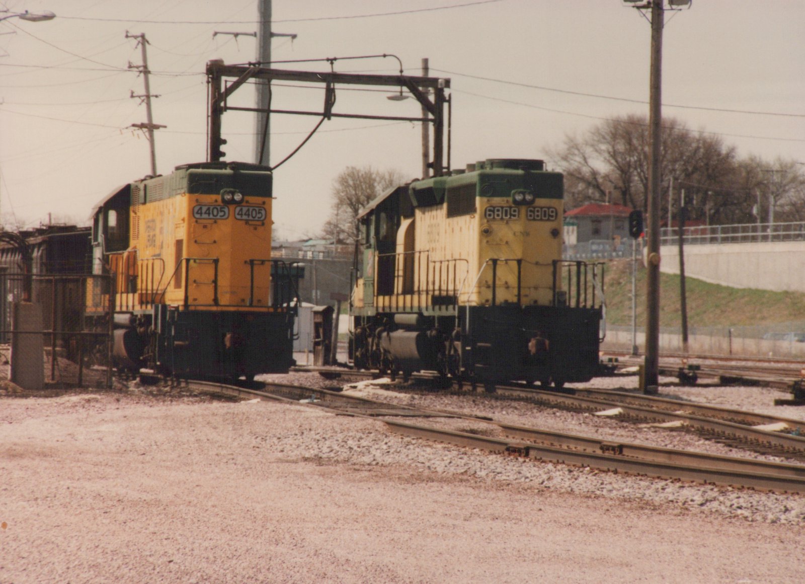 Two yellow and green CNW diesel locomotives, 4405 and 6809, are parked on parallel tracks in a gravel yard under an overcast sky.
