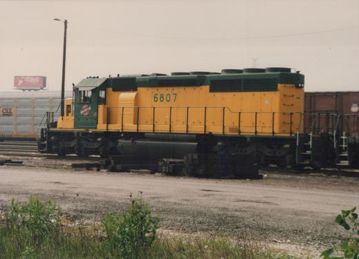 A side view of a yellow and dark green diesel locomotive numbered 6807, with 'NORTHWESTERN' lettering on the cab, standing on a track in a gravel yard with freight cars and a billboard in the background.