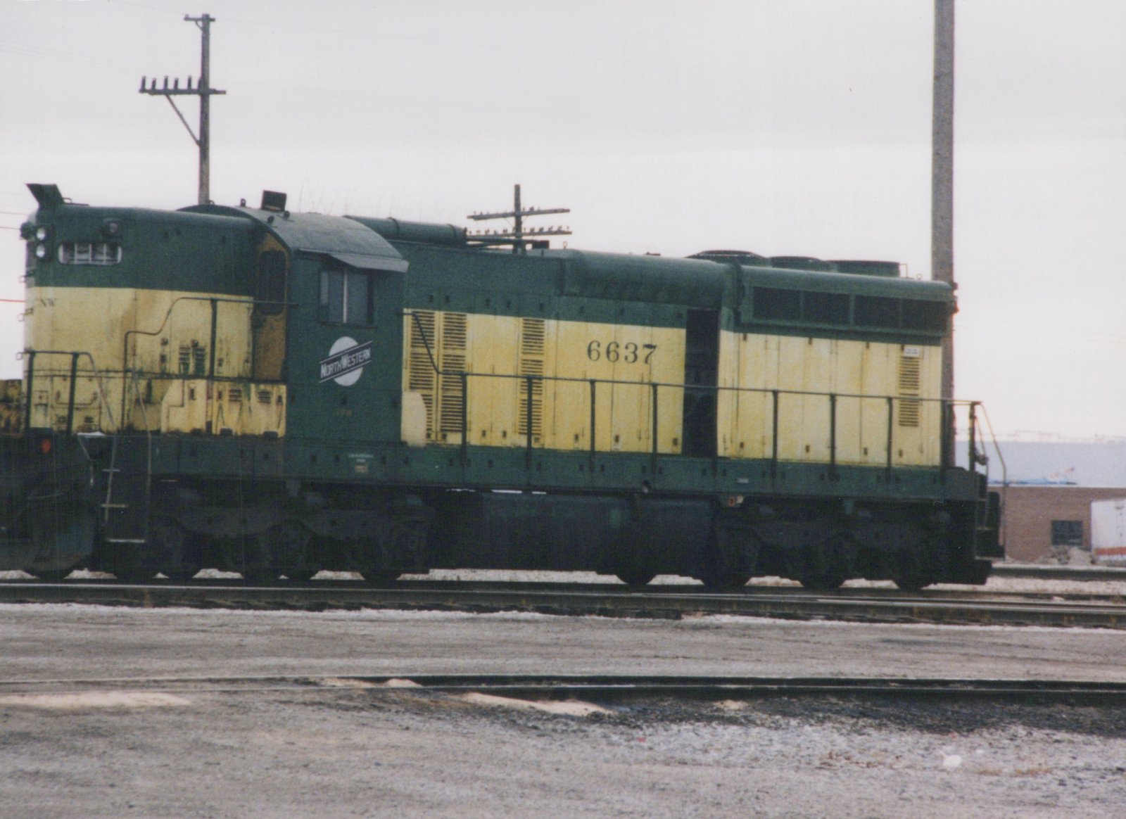 A green and yellow diesel locomotive with the number 6637 and 'NORTHWESTERN' lettering sits on tracks in a snowy yard under an overcast sky.