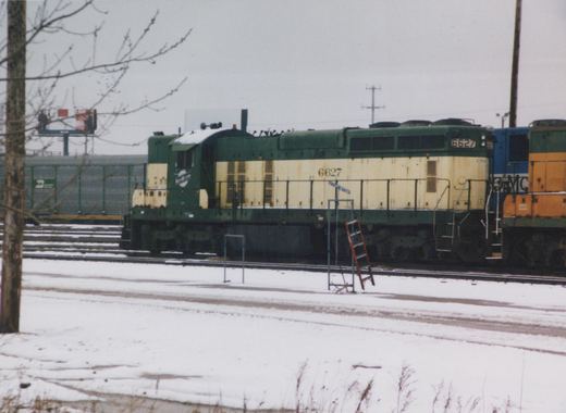 Side view of a green and cream Chicago and North Western diesel locomotive, number 6627, parked in a snowy rail yard with another locomotive and freight cars in the background.