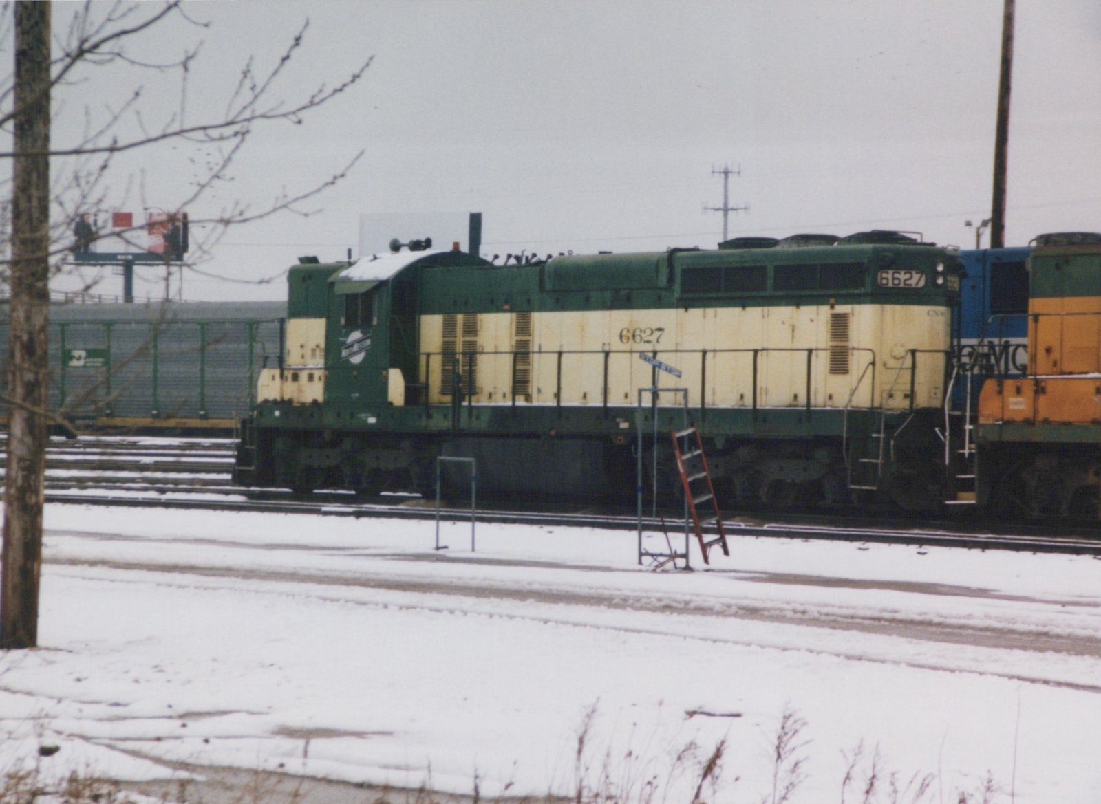 Side view of a green and cream Chicago and North Western diesel locomotive, number 6627, parked in a snowy rail yard with another locomotive and freight cars in the background.