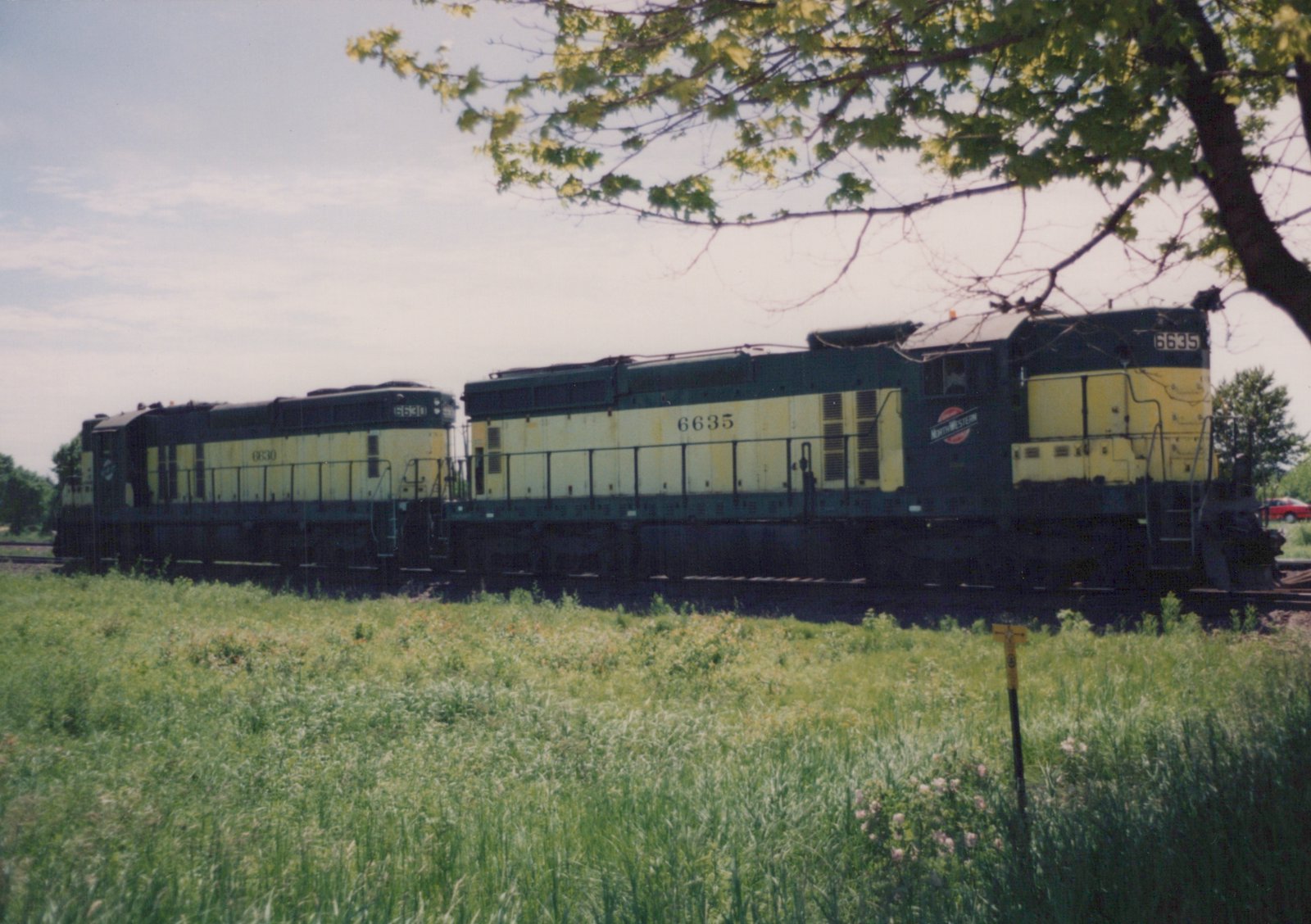 Two dark green and yellow diesel locomotives with numbers 6635 and 6630, marked Chicago and North Western, are parked on a railroad track next to a field of tall grass under a tree.
