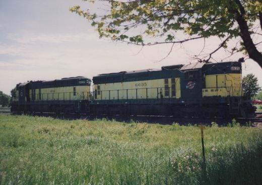 Two dark green and yellow diesel locomotives with numbers 6635 and 6630, marked Chicago and North Western, are parked on a railroad track next to a field of tall grass under a tree.