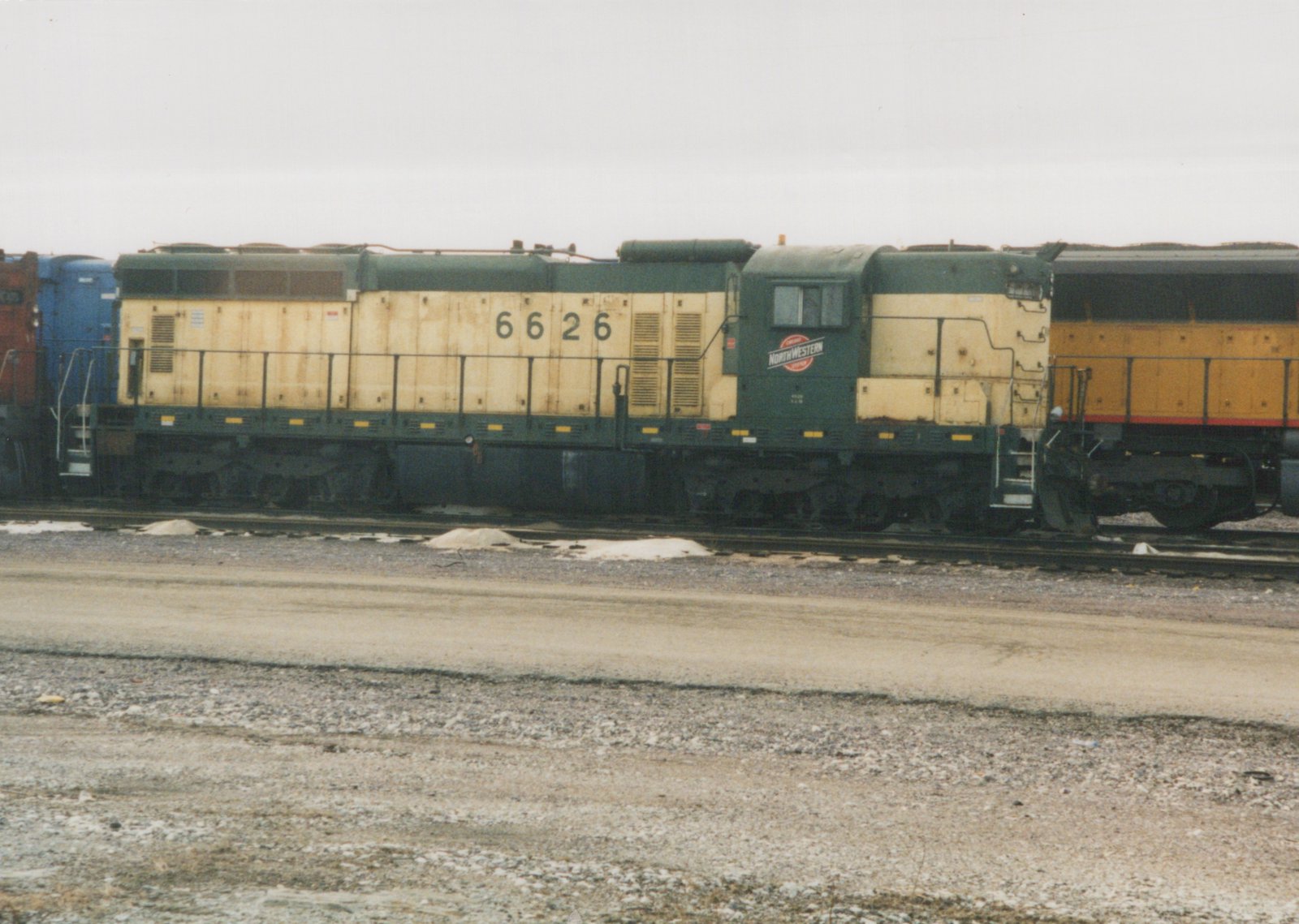 A side view of Chicago and North Western diesel locomotive number 6626, painted in green and yellow, parked on tracks in a rail yard with snow on the ground.