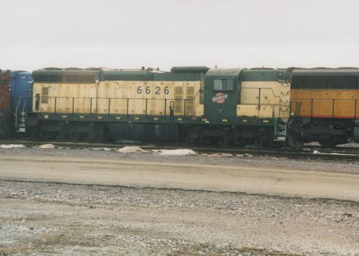 A side view of Chicago and North Western diesel locomotive number 6626, painted in green and yellow, parked on tracks in a rail yard with snow on the ground.