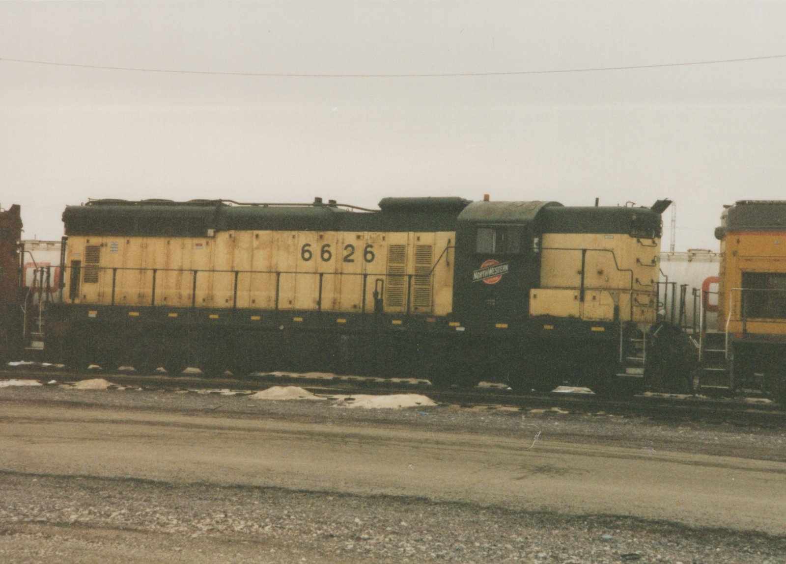 A side view of a yellow and black Chicago and North Western diesel locomotive, number 6626, parked in a rail yard with snow patches on the ground under an overcast sky.