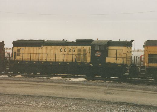 A side view of a yellow and black Chicago and North Western diesel locomotive, number 6626, parked in a rail yard with snow patches on the ground under an overcast sky.
