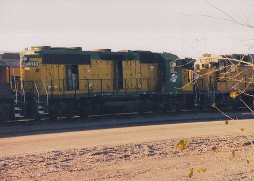 A yellow and green Chicago and North Western diesel locomotive, number 5535, is parked in a rail yard with bare branches in the foreground.