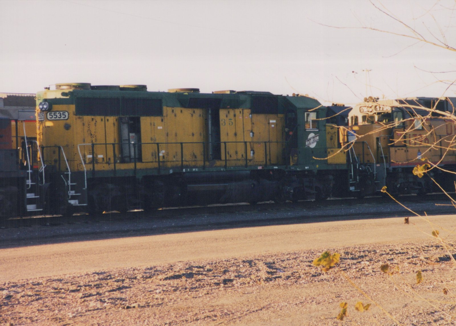 A yellow and green Chicago and North Western diesel locomotive, number 5535, is parked in a rail yard with bare branches in the foreground.