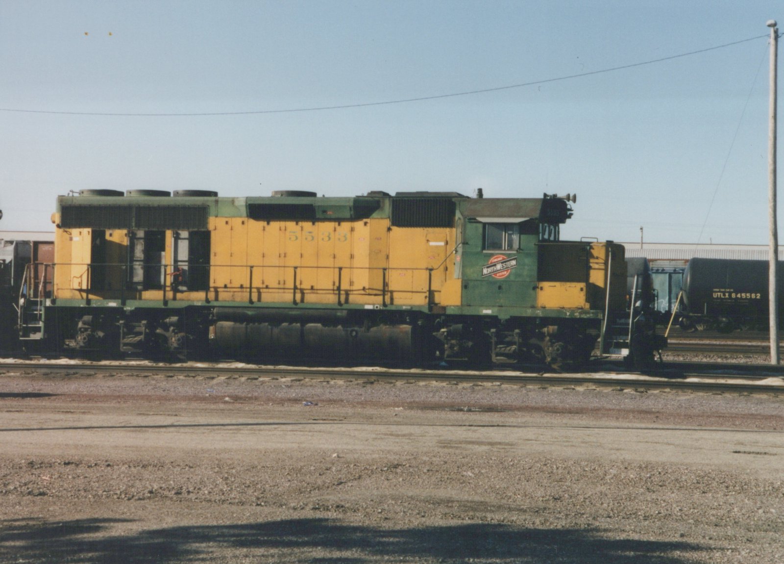 A side view of a yellow and green Chicago and North Western diesel locomotive with the number 5533, standing on tracks in a gravel yard.