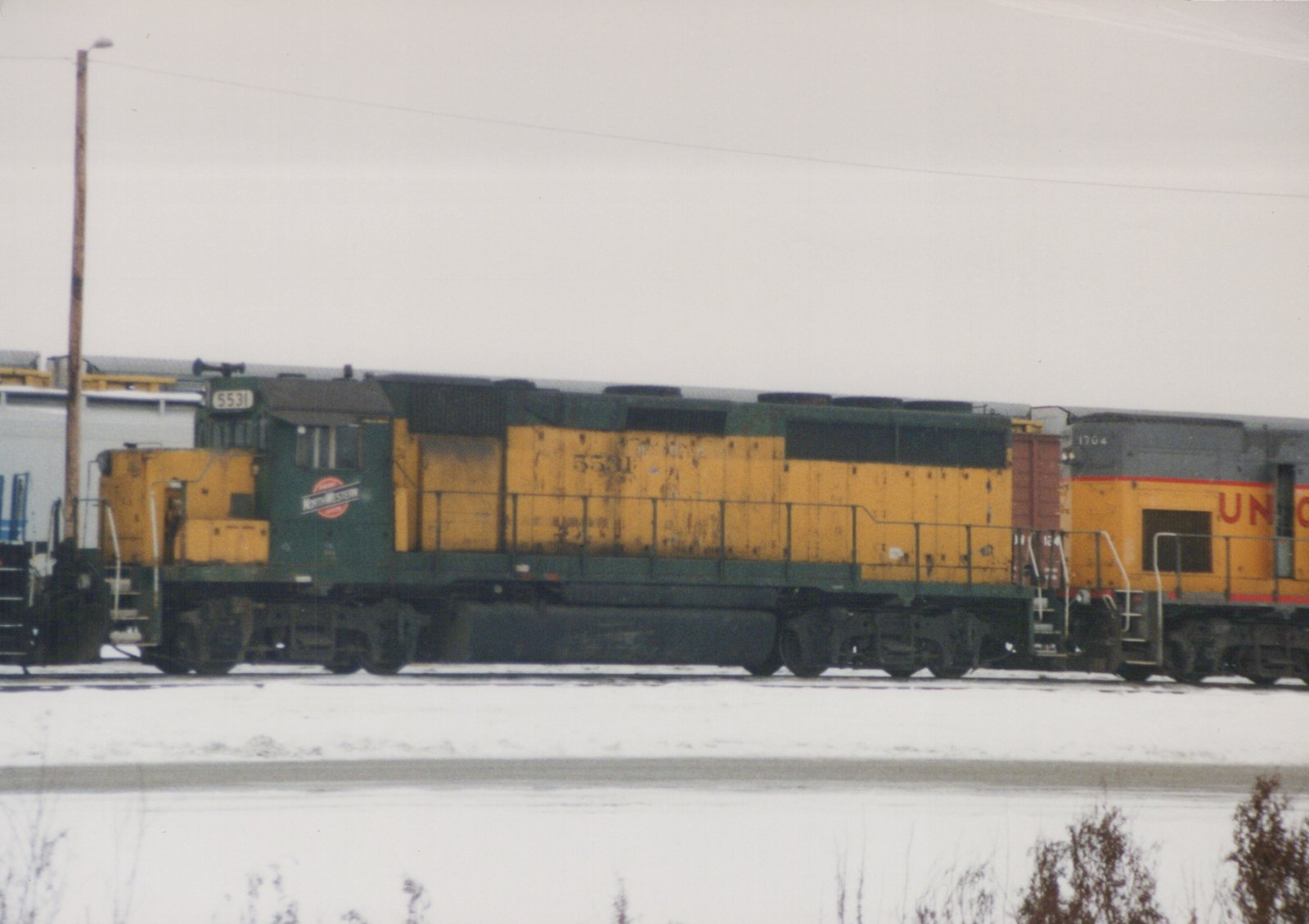 Side view of a green and yellow Chicago and North Western GP9 locomotive number 5531 parked in a snowy yard, with a yellow and gray Union Pacific locomotive number 1704 partially visible behind it.