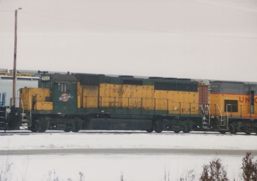 Side view of a green and yellow Chicago and North Western GP9 locomotive number 5531 parked in a snowy yard, with a yellow and gray Union Pacific locomotive number 1704 partially visible behind it.