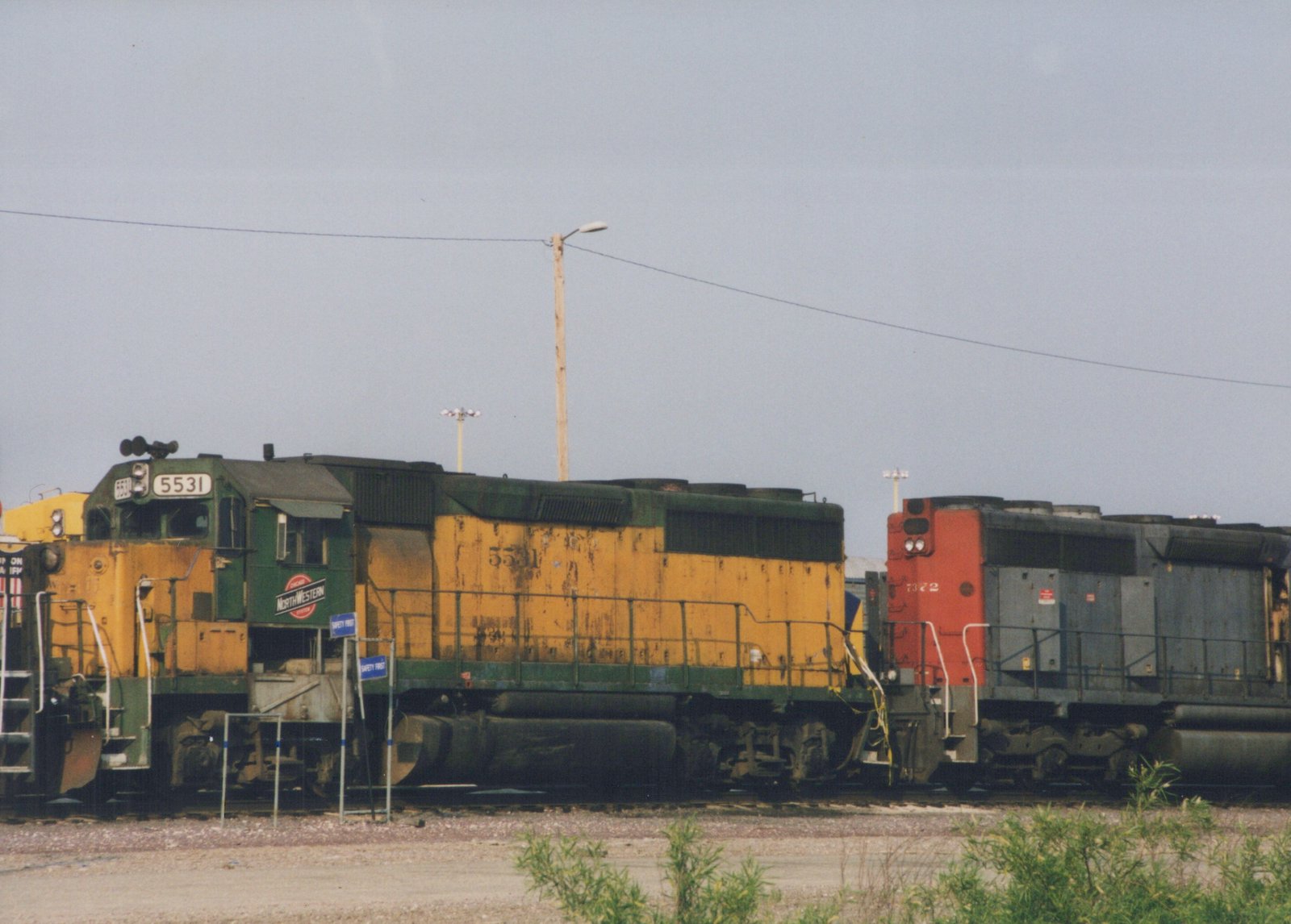 Side view of two diesel locomotives in a railroad yard on an overcast day; the lead locomotive is green and yellow with the number 5531, and the trailing locomotive is red and gray with the number 7322.