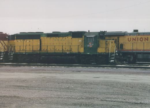Side view of a yellow and green Chicago and North Western diesel locomotive, number 5533, parked on tracks in a yard with a Union Pacific locomotive behind it under an overcast sky.