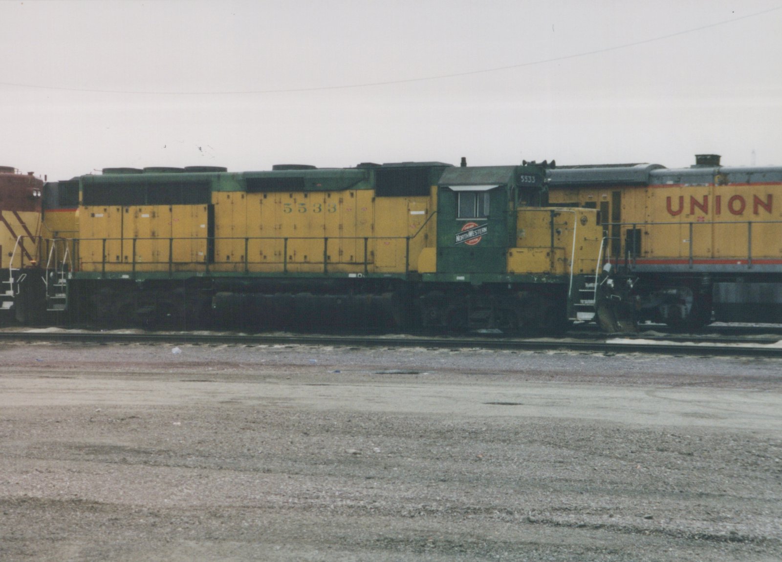 Side view of a yellow and green Chicago and North Western diesel locomotive, number 5533, parked on tracks in a yard with a Union Pacific locomotive behind it under an overcast sky.