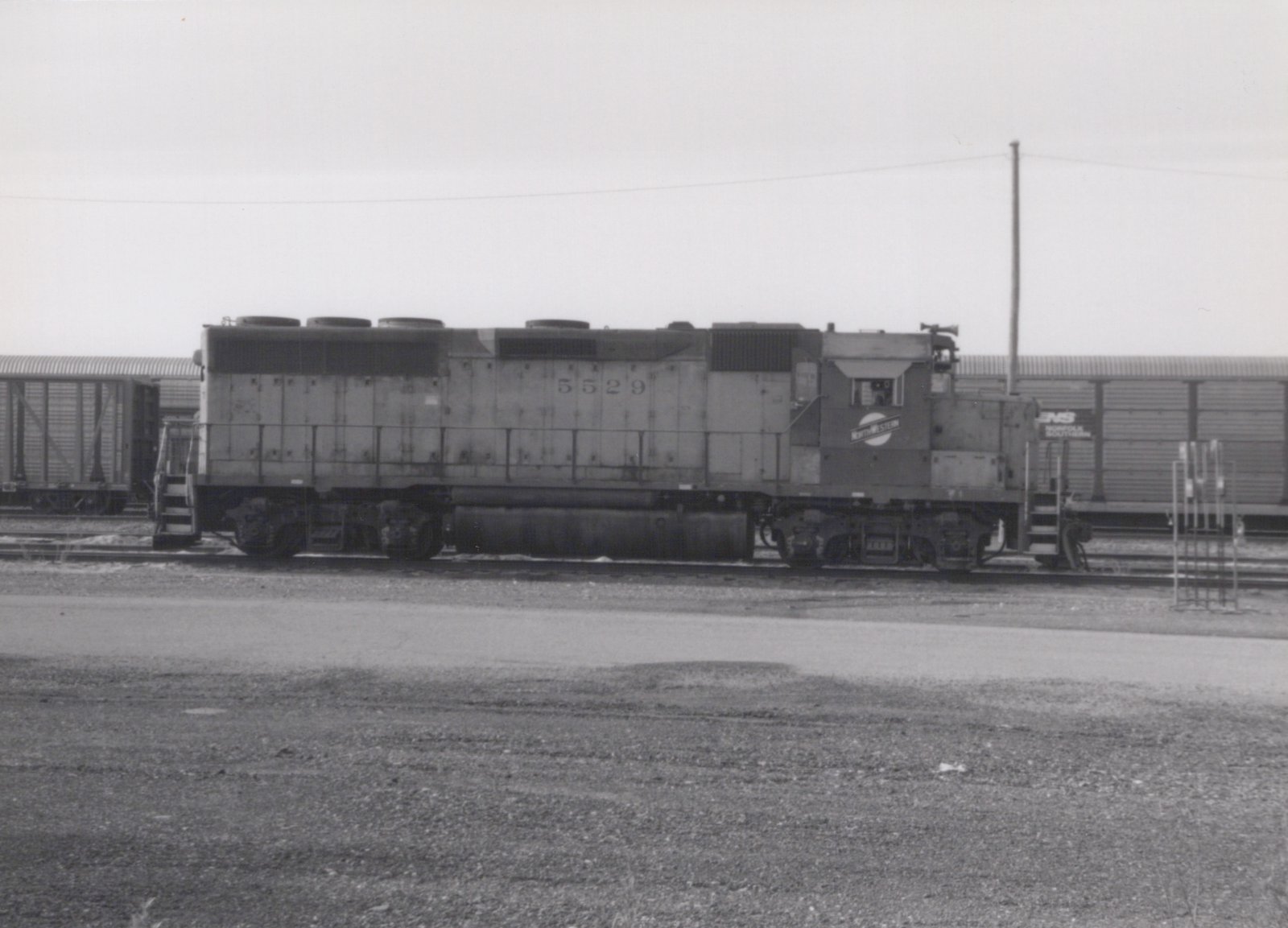 A black and white photograph of a Chicago and North Western diesel locomotive numbered 5529, seen in side profile in a rail yard with freight cars and a gravel foreground.