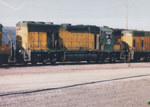 A yellow and green Chicago and North Western diesel locomotive, number 5529, is parked in a gravel yard with another locomotive partially visible behind it.