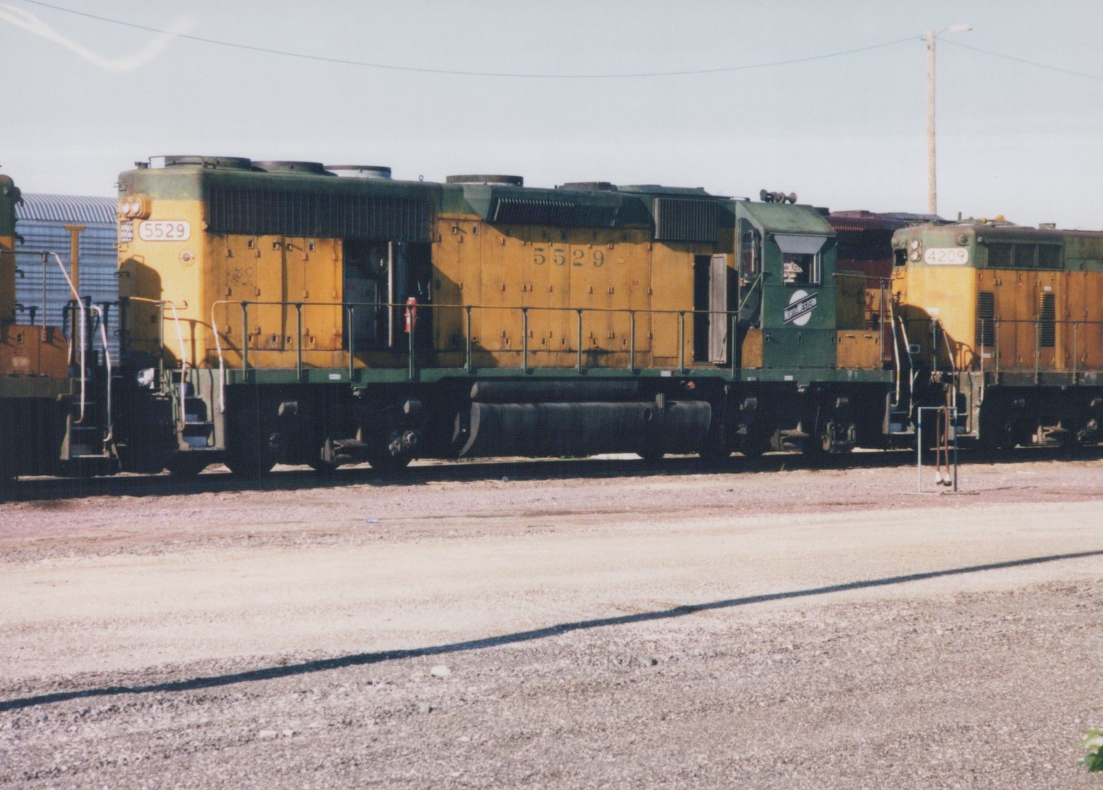 A yellow and green Chicago and North Western diesel locomotive, number 5529, is parked in a gravel yard with another locomotive partially visible behind it.