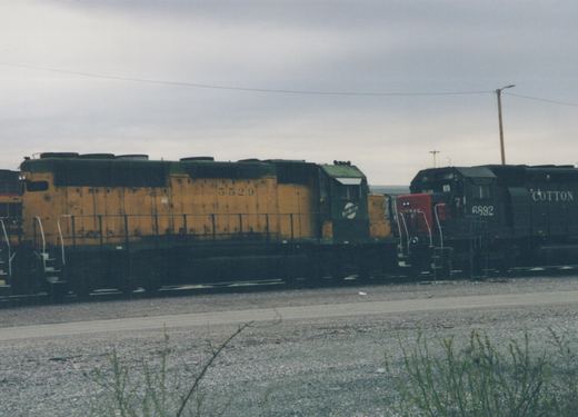 Two diesel locomotives, one yellow and green with the number 5529 and the name 'North Western', and one black with the number 6892 and the name 'Cotton', are parked side by side on railroad tracks in a gravel yard under a cloudy sky.