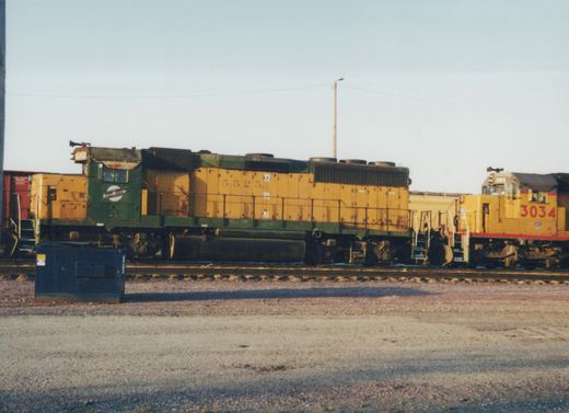 A side view of two yellow and green Chicago and North Western diesel locomotives, numbers 5525 and 3034, sitting on tracks in a gravel yard under a clear sky.