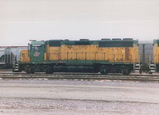 A green and yellow Chicago and North Western diesel locomotive numbered 5520 is parked on tracks in a rail yard under an overcast sky, with another similar locomotive visible behind it.