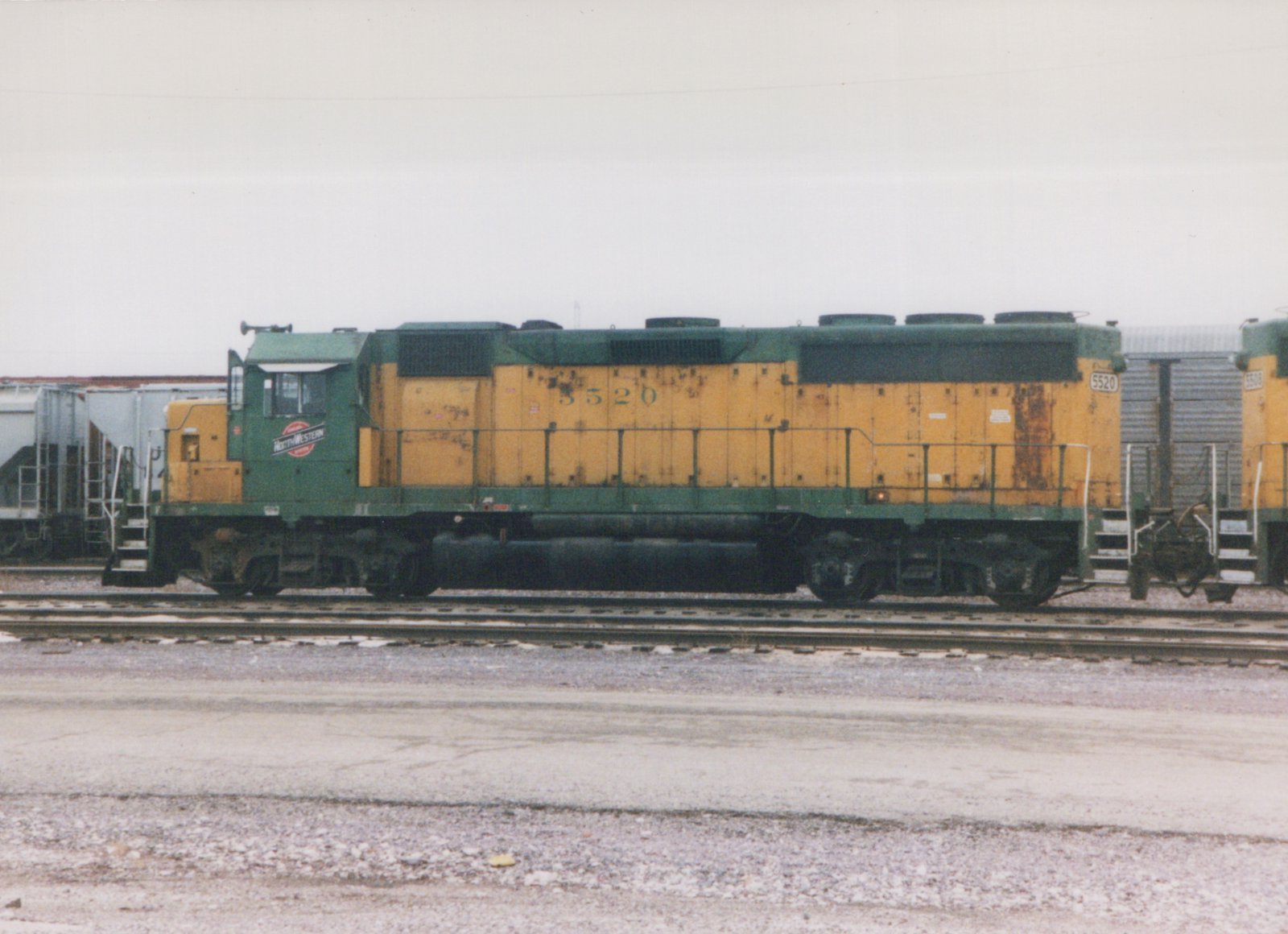 A green and yellow Chicago and North Western diesel locomotive numbered 5520 is parked on tracks in a rail yard under an overcast sky, with another similar locomotive visible behind it.
