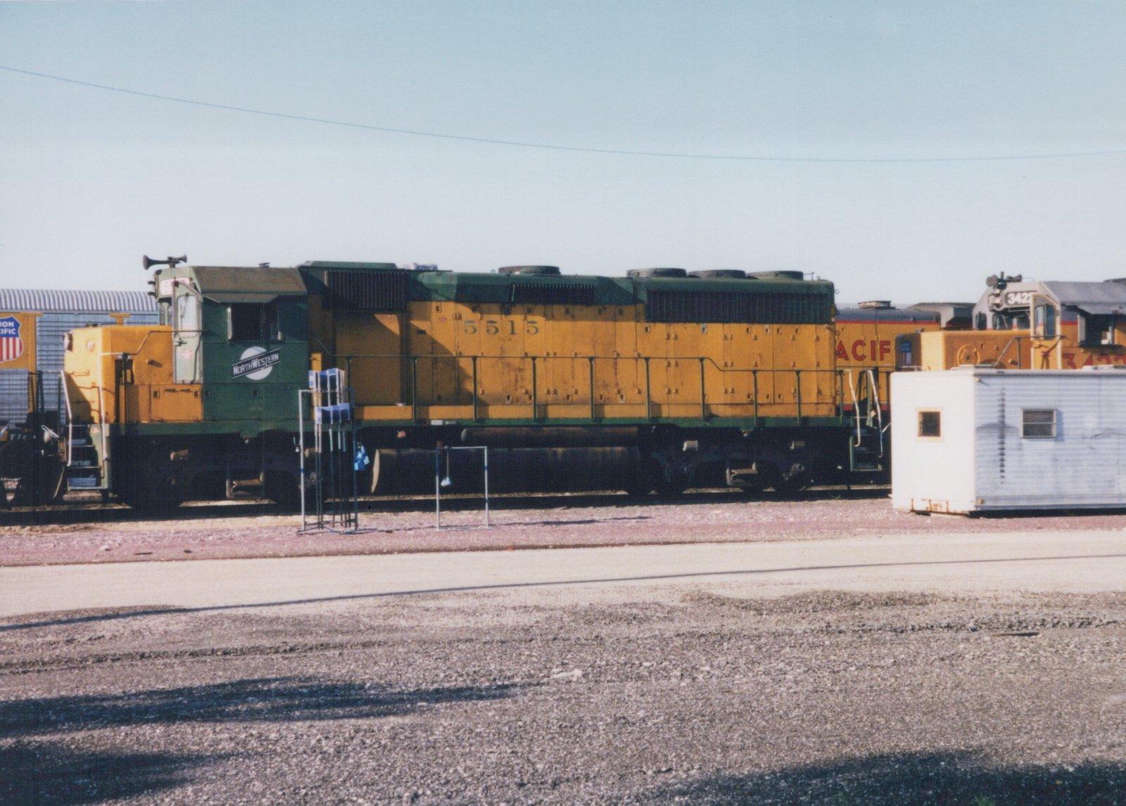 Side view of a yellow and green Chicago and North Western diesel locomotive numbered 5515, parked in a gravel rail yard with another locomotive visible in the background.