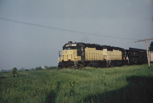 A two-unit diesel locomotive painted in yellow and dark green with the letters CNW on the side moves along a track next to a green, grassy field under an overcast sky.