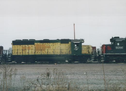 Side view of a yellow and green Chicago and North Western locomotive number 5512 parked in a gravel yard with another locomotive partially visible in the background.