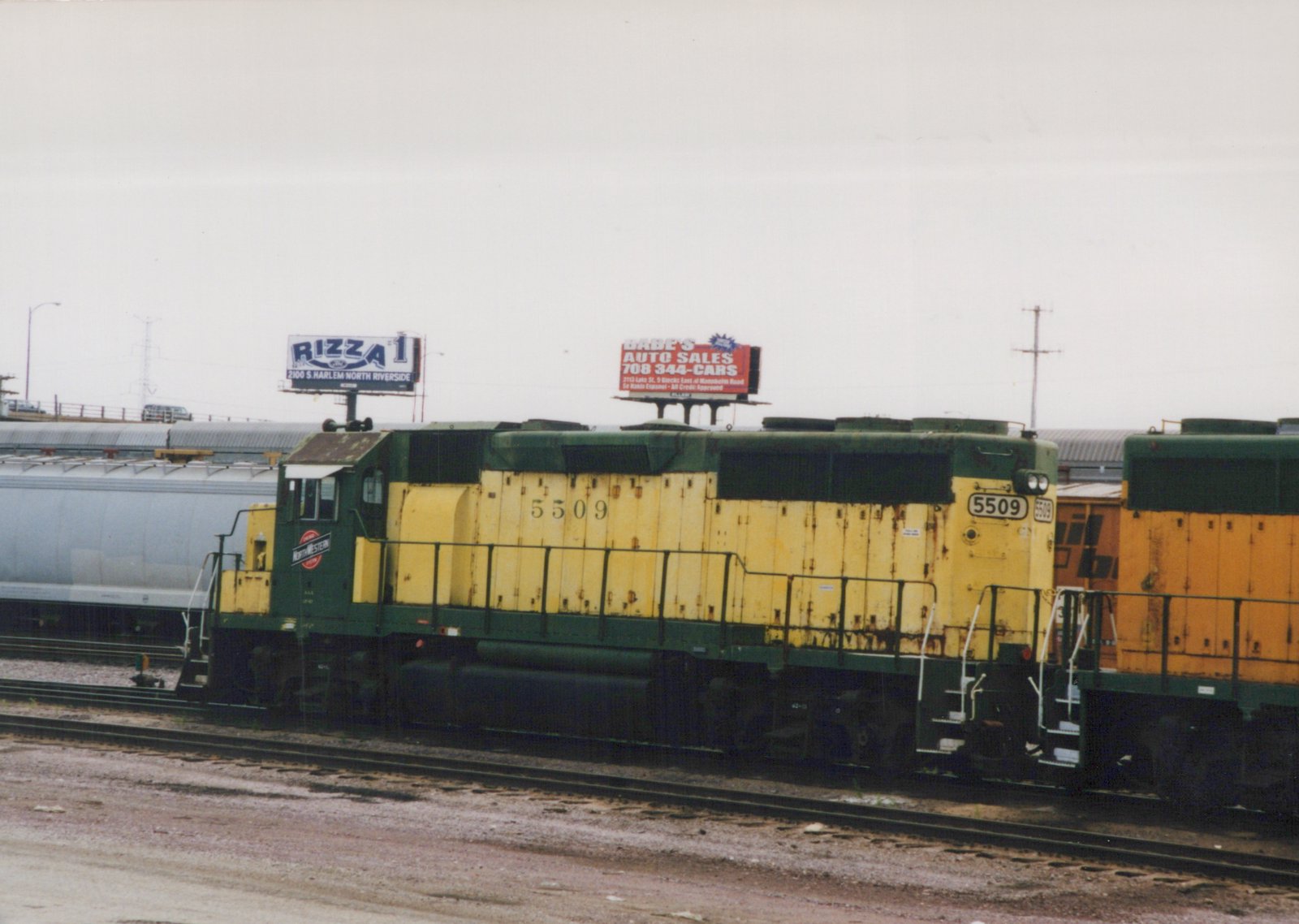 A green and yellow diesel locomotive with the number 5509 and a North Western herald sits on railroad tracks in a yard, with billboards visible in the background under an overcast sky.
